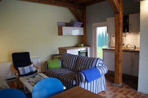 Uma sala de estar aconchegante com um sofá listrado, uma cadeira de madeira e uma mesa de jantar. Uma cozinha é visível ao fundo, com armários brancos e uma janela.