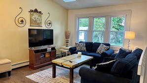 A living room with a TV, sofa, coffee table, and a window with a view of trees. -  Great 5 Bedroom in Beautiful Bonnet Shores, Narragansett R (Narragansett)