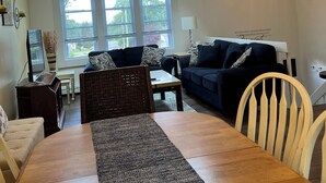 A dining area with a wooden table, chairs, and a ceiling fan. There are two windows with blinds, a sofa, and a TV on a stand. -  Great 5 Bedroom in Beautiful Bonnet Shores, Narragansett R (Narragansett)