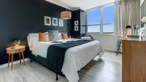 Une chambre avec une grande fenêtre, un lit avec une literie blanche, une table de chevet avec une lampe et une commode.