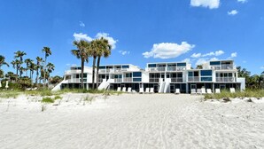 Ein Strandhotel mit mehreren Balkonen, einem Sandstrand davor und Palmen.