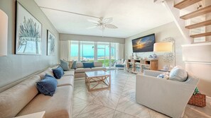 A spacious living room with a large sofa, a coffee table, and a striped armchair. There is a ceiling fan, a painting, and a view of the ocean through large windows.