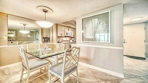 A dining area with a glass table and chairs, a kitchen in the background, and a large decorative piece on the wall.