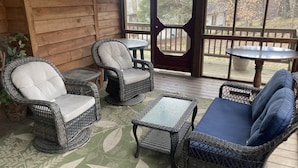 A covered outdoor seating area with wicker furniture and a glass table, featuring a wooden deck and a view of trees.