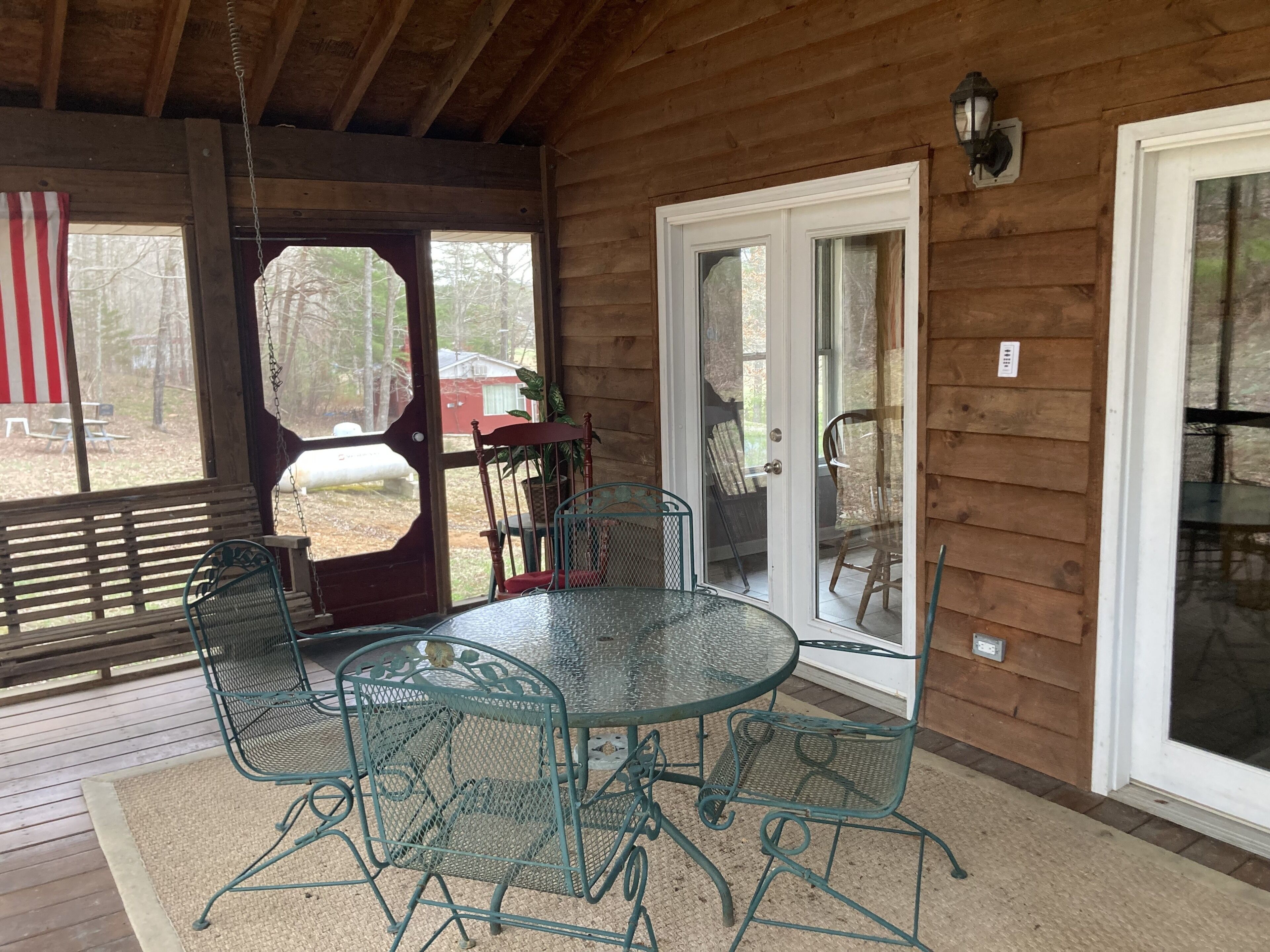 A covered patio with a round glass table and metal chairs, wooden walls, and a sliding glass door leading outside.
