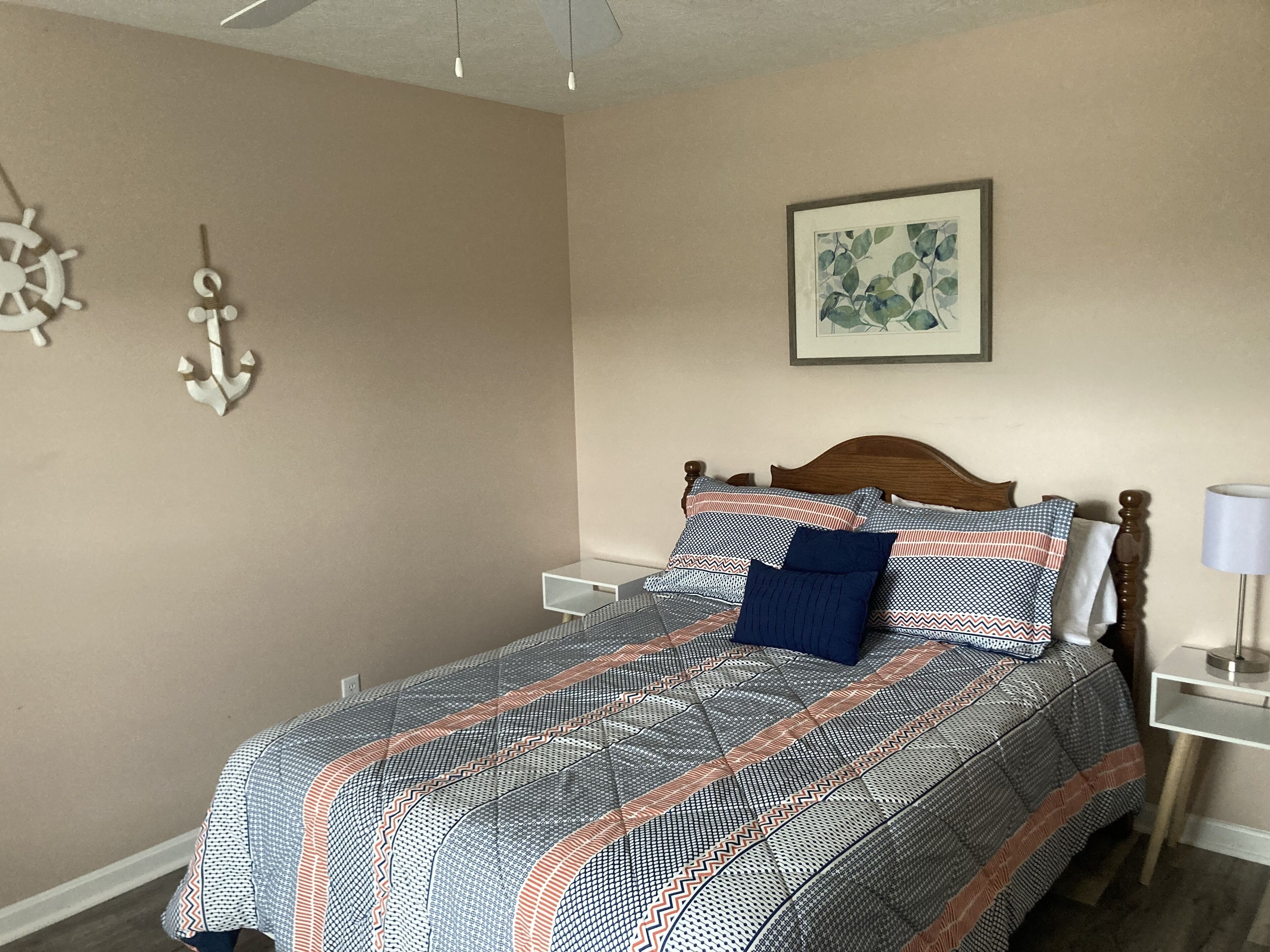 A neatly made bed with patterned bedding, a wooden headboard, a bedside table with a lamp, and wall decorations including a ship wheel and an anchor.