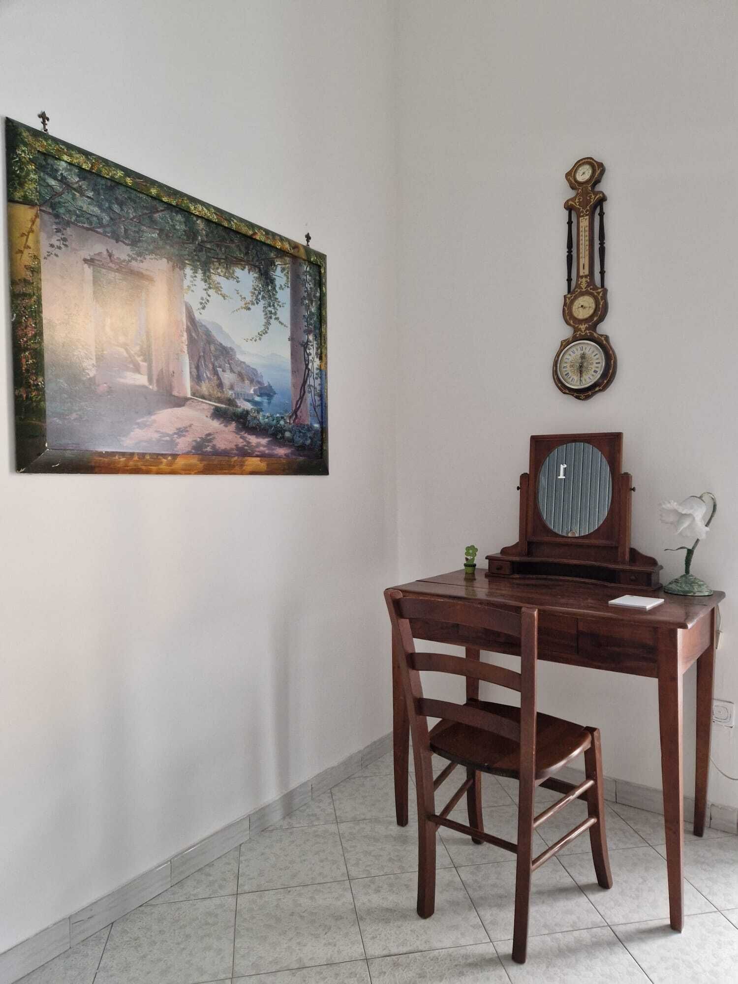 Quarto com escrivaninha de madeira, cadeira, quadro emoldurado e relógio fixado na parede.