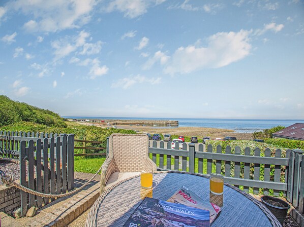 Una silla de mimbre con cojín, una cerveza y una revista de viajes sobre una mesa con vistas al mar y la playa.