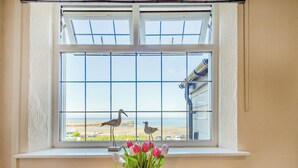 A window with a view of the beach and two seagulls, a vase with flowers, and a table with a glass and a plate.