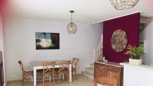 A dining area with a white table and chairs, a wicker chair with a cushion, a pendant light, and a wall clock. - 3-star listed house with south-facing courtyard in La noue (Sainte-Marie-de-Ré)