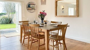 A dining area with a wooden table, chairs, a vase with flowers, and a wall-mounted shelf. - 3 bedroom accommodation in St Mawgan (St Mawgan)