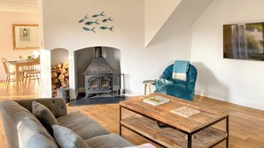 A living room with a sofa, a coffee table, a fireplace, a blue armchair, and a dining area with chairs and a table. - 3 bedroom accommodation in St Mawgan (St Mawgan)