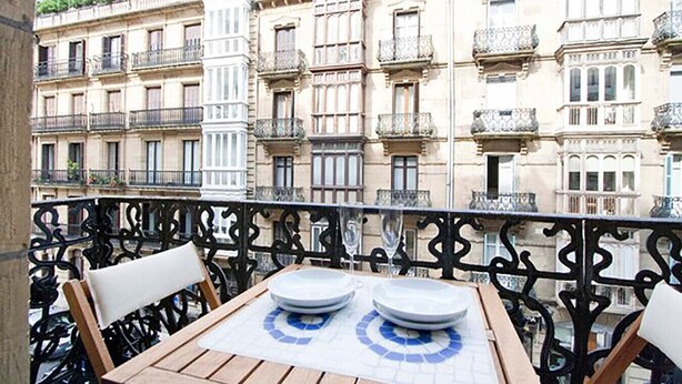 Un balcon avec une table dressée pour deux personnes, offrant une vue sur une rangée de bâtiments de style européen classique, chacun doté d’un balcon.