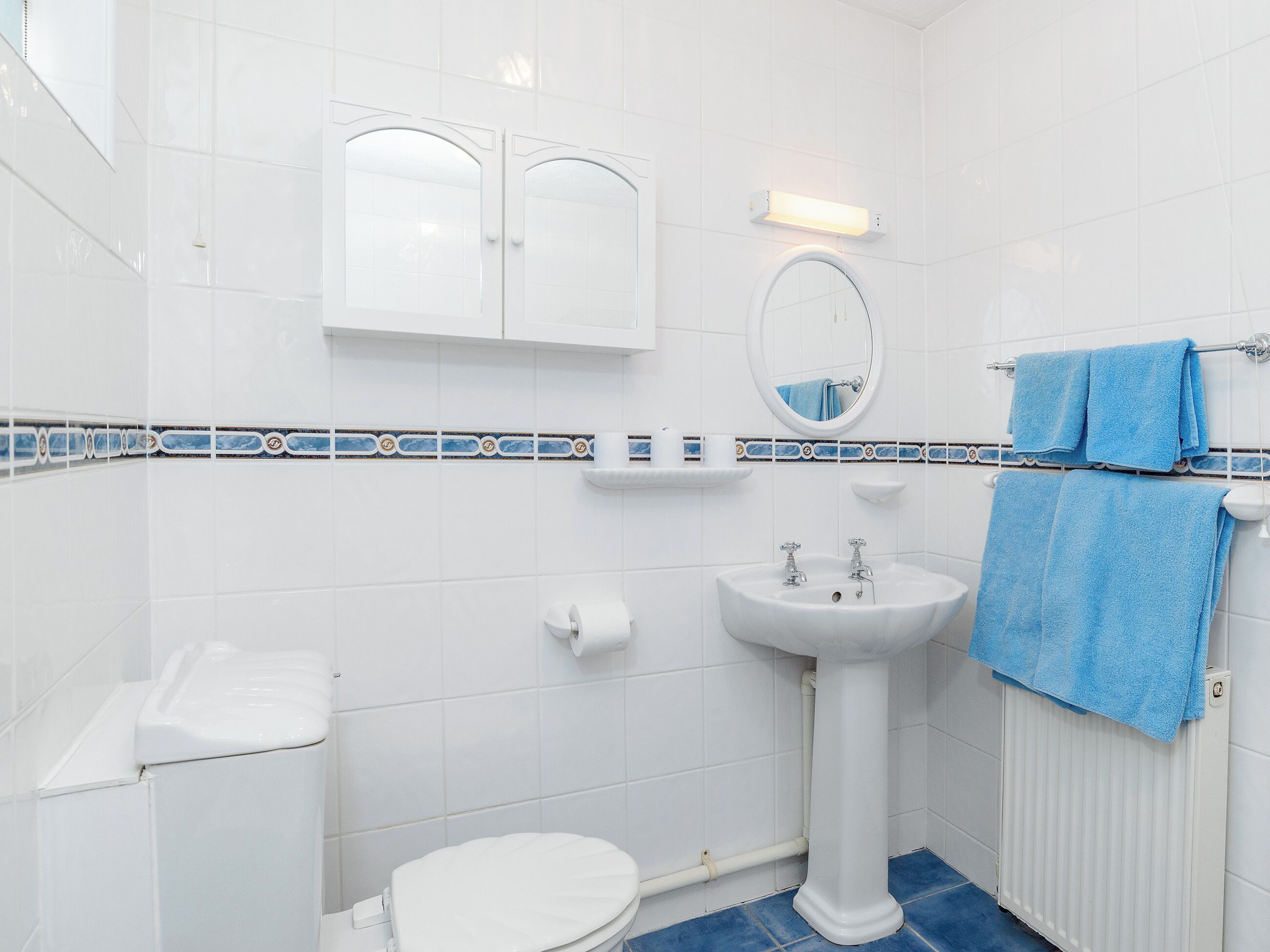 Ein Badezimmer mit weißen Fliesen, einem Standwaschbecken, einer Toilette, einem Spiegel und einem Handtuchhalter mit blauen Handtüchern.