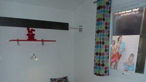 A room with two single beds, a red shelf, a window with a polka dot curtain, and a poster on the wall.