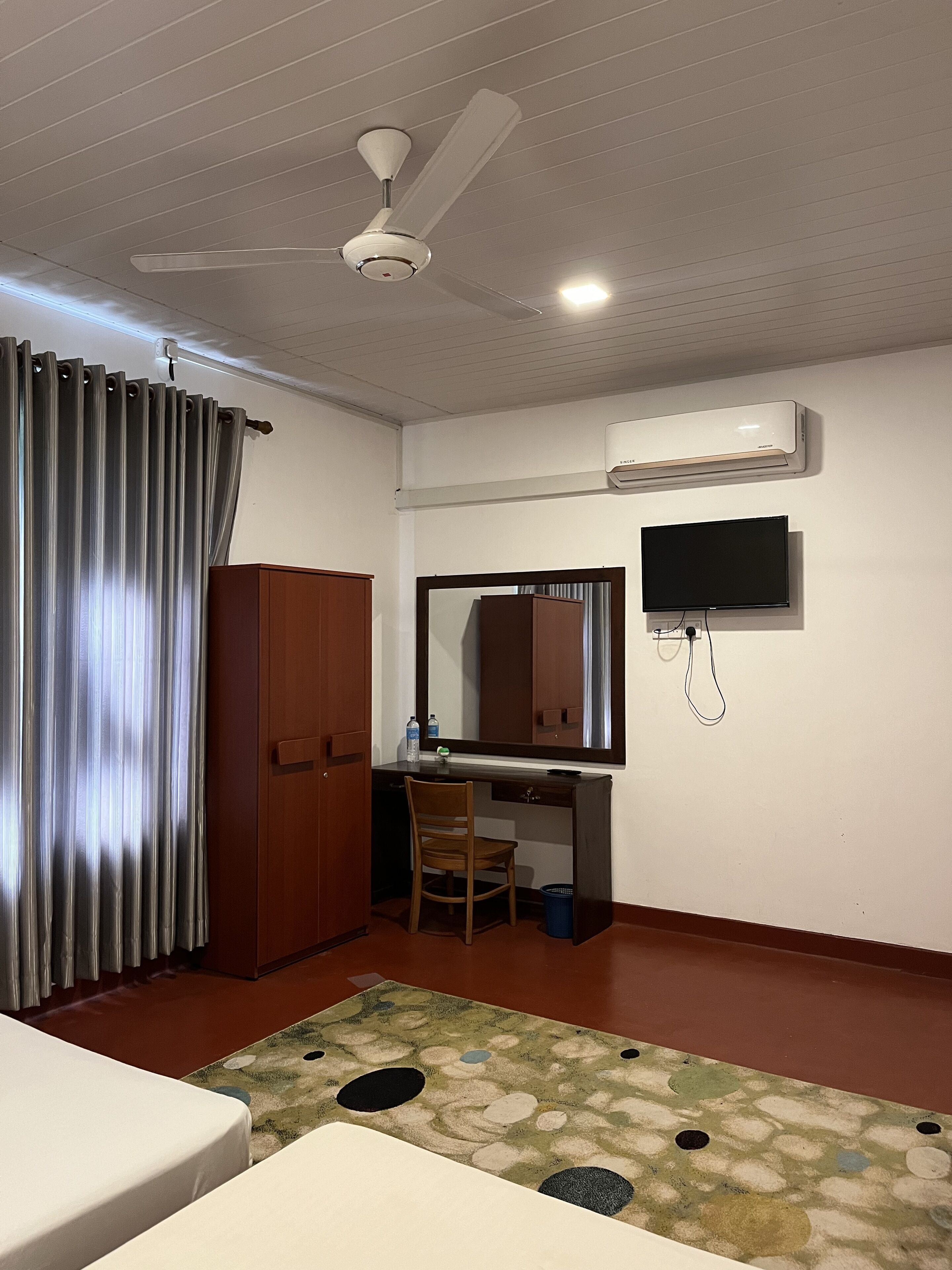A hotel room with a bed, a desk with a chair, a ceiling fan, an air conditioner, and a television.