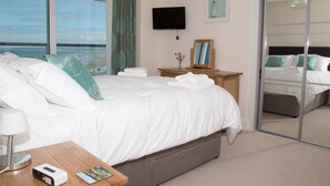 Habitación de hotel con una cama grande, televisión, espejo, mesita de noche con lámpara y vista al mar.