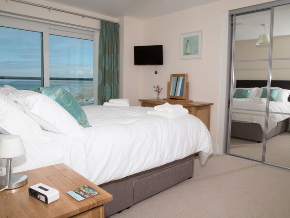 A hotel room with a large bed, a TV, a mirror, a bedside table with a lamp, and a view of the sea.
