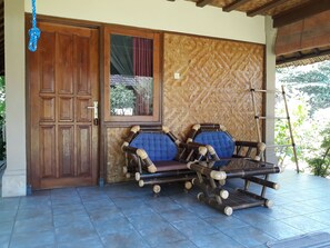 A wooden door, a window with a view, a woven wall, and two blue cushioned chairs on a tiled floor. - Le Jardin (Karangasem)
