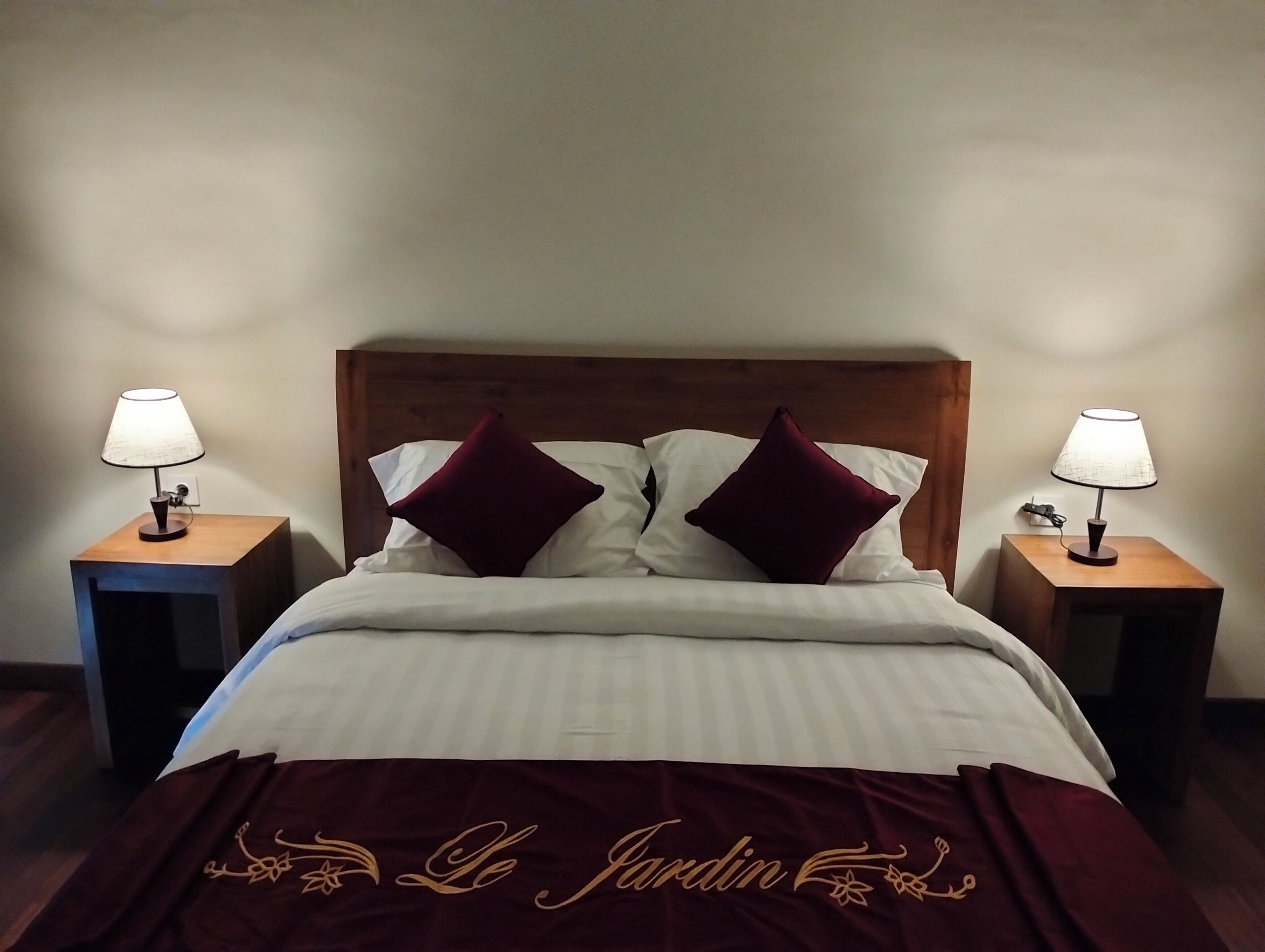Un lit bien fait, recouvert d’une couvre-lit rouge foncé, avec deux oreillers blancs et deux coussins décoratifs rouges assortis. Il y a deux tables de chevet avec des lampes, et la chambre est dotée d’une tête de lit en bois.