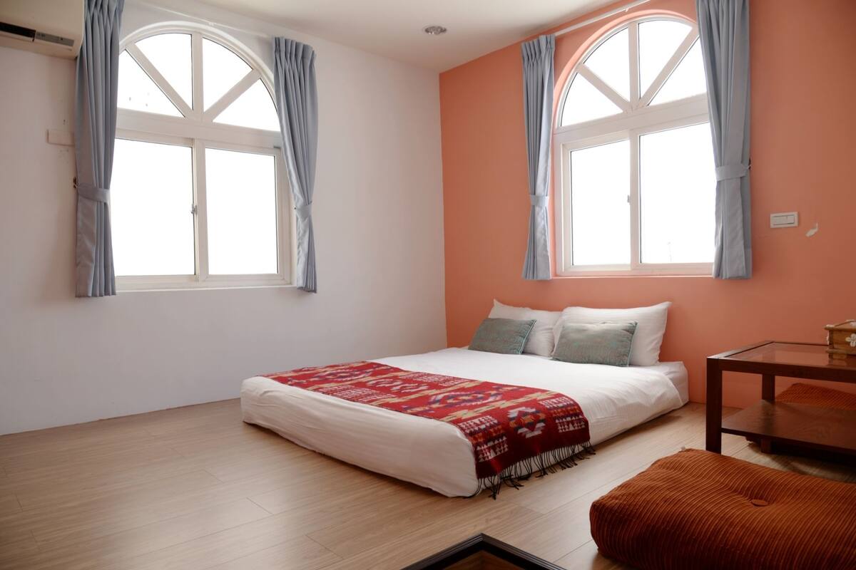 A bedroom with a bed, two windows with curtains, a wooden floor, and a small table with a chair.
