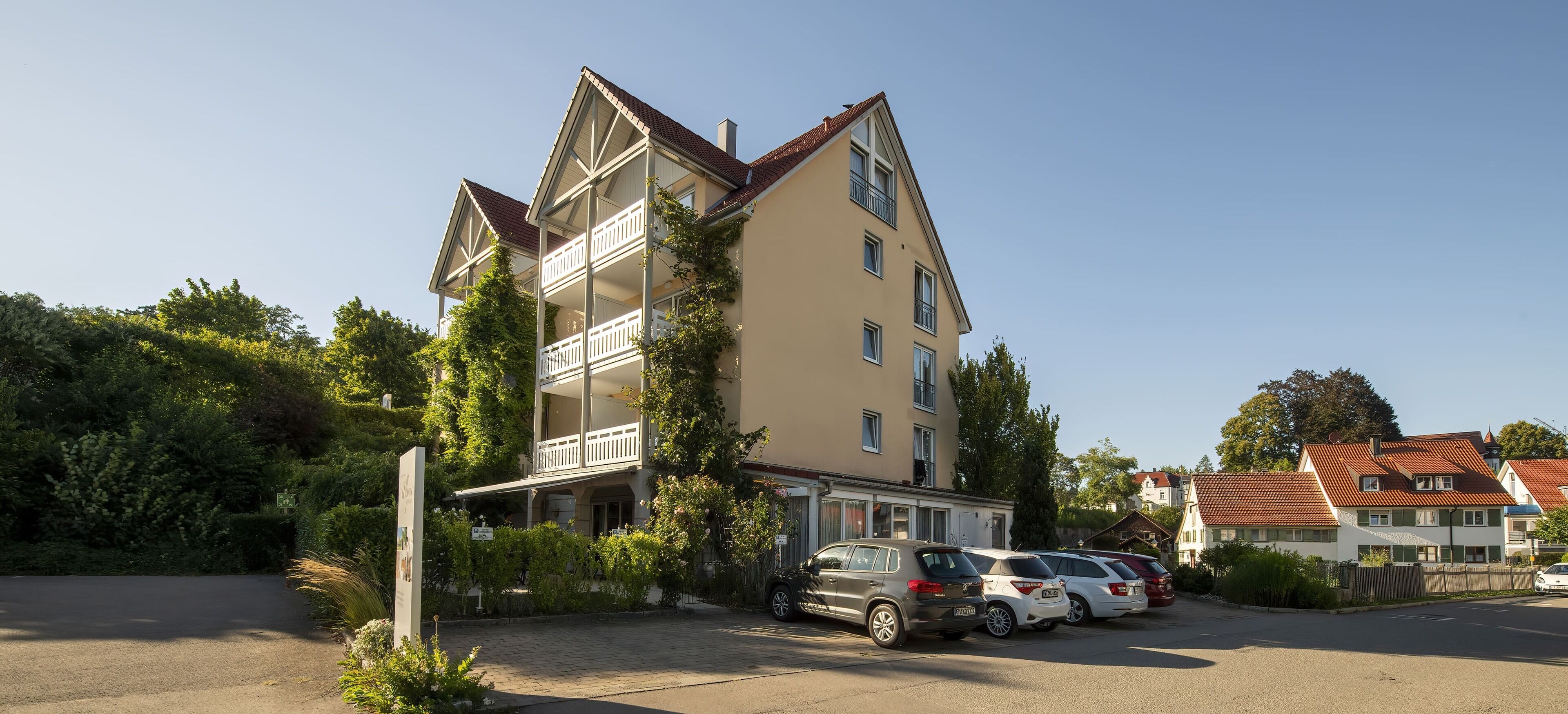 A multi-story building with a balcony, parked cars, and greenery.