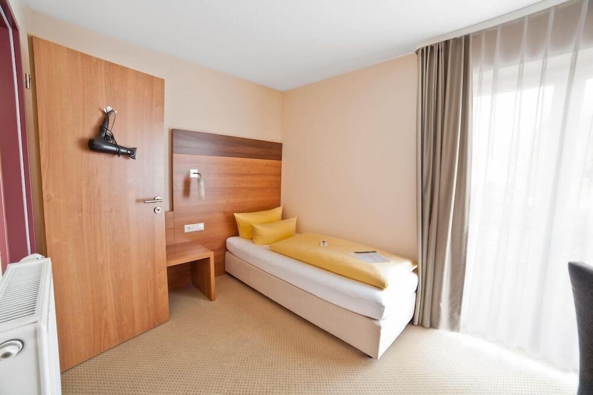 Ein kleines Hotelzimmer mit einem Einzelbett, einem Holzschrank, einem kleinen Schreibtisch und einem Fenster mit Vorhängen.