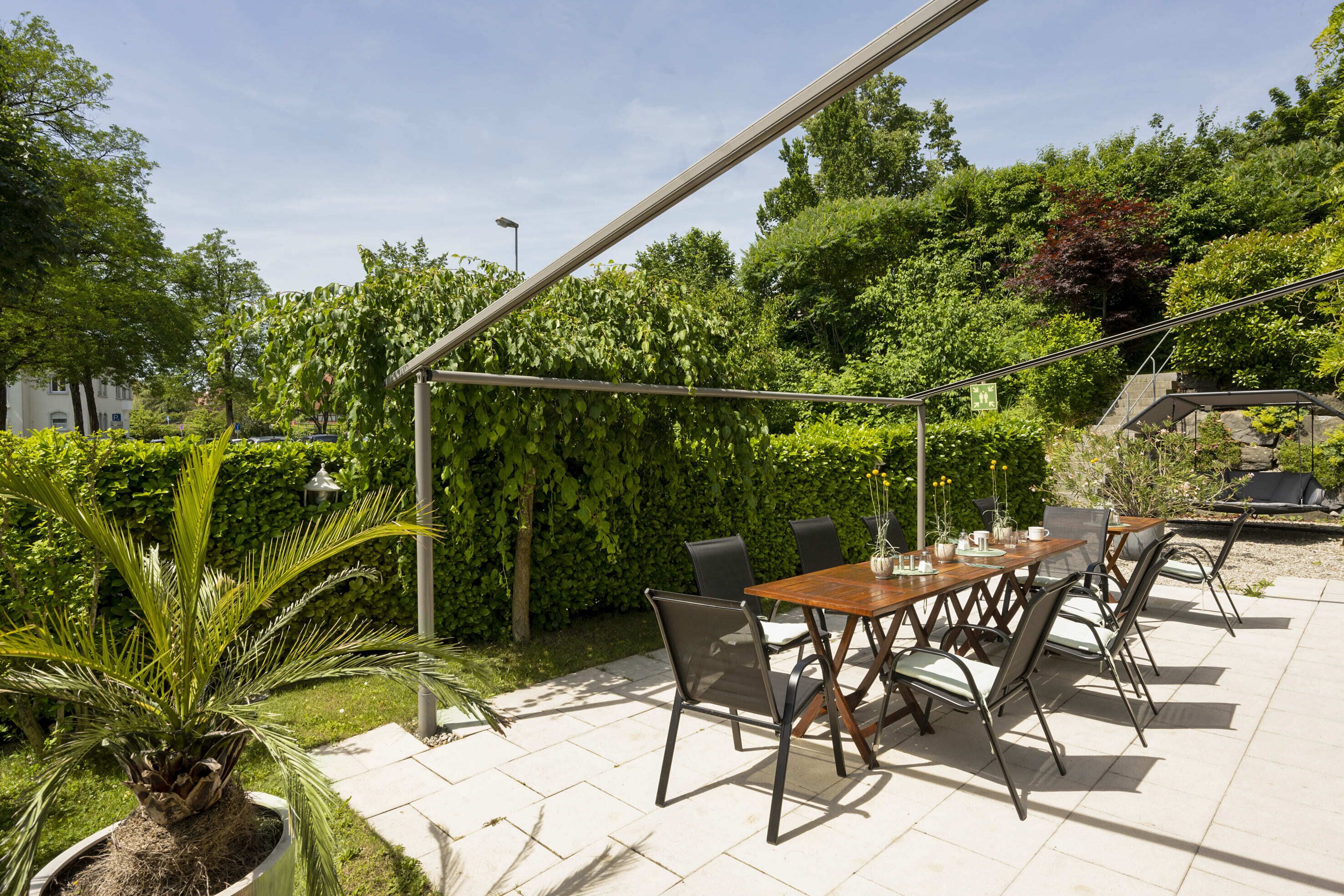 An outdoor patio with a wooden table and chairs, surrounded by greenery and a pergola.