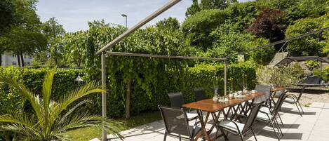 An outdoor patio with a wooden table and chairs, surrounded by greenery and a pergola.