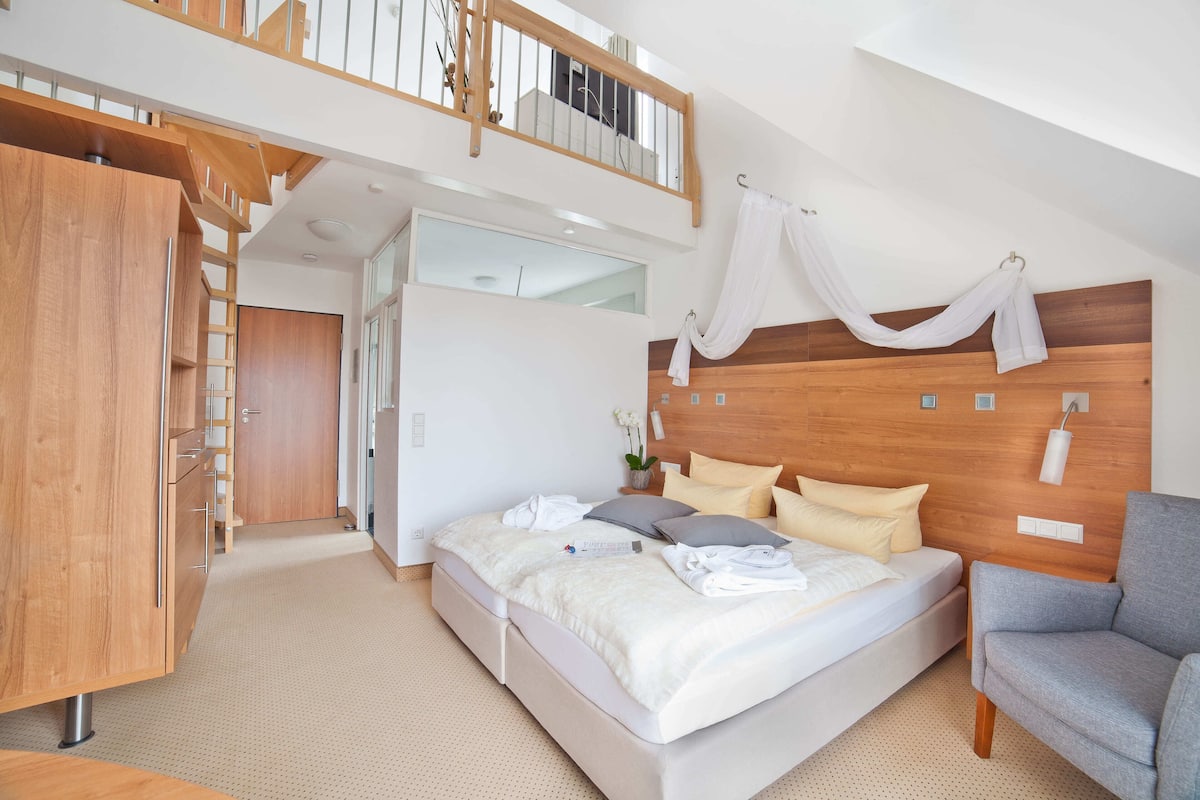 Ein modernes Schlafzimmer mit Schlafplatz in Podesteinlage, einem Holzkopfende und einem grauen Sessel.