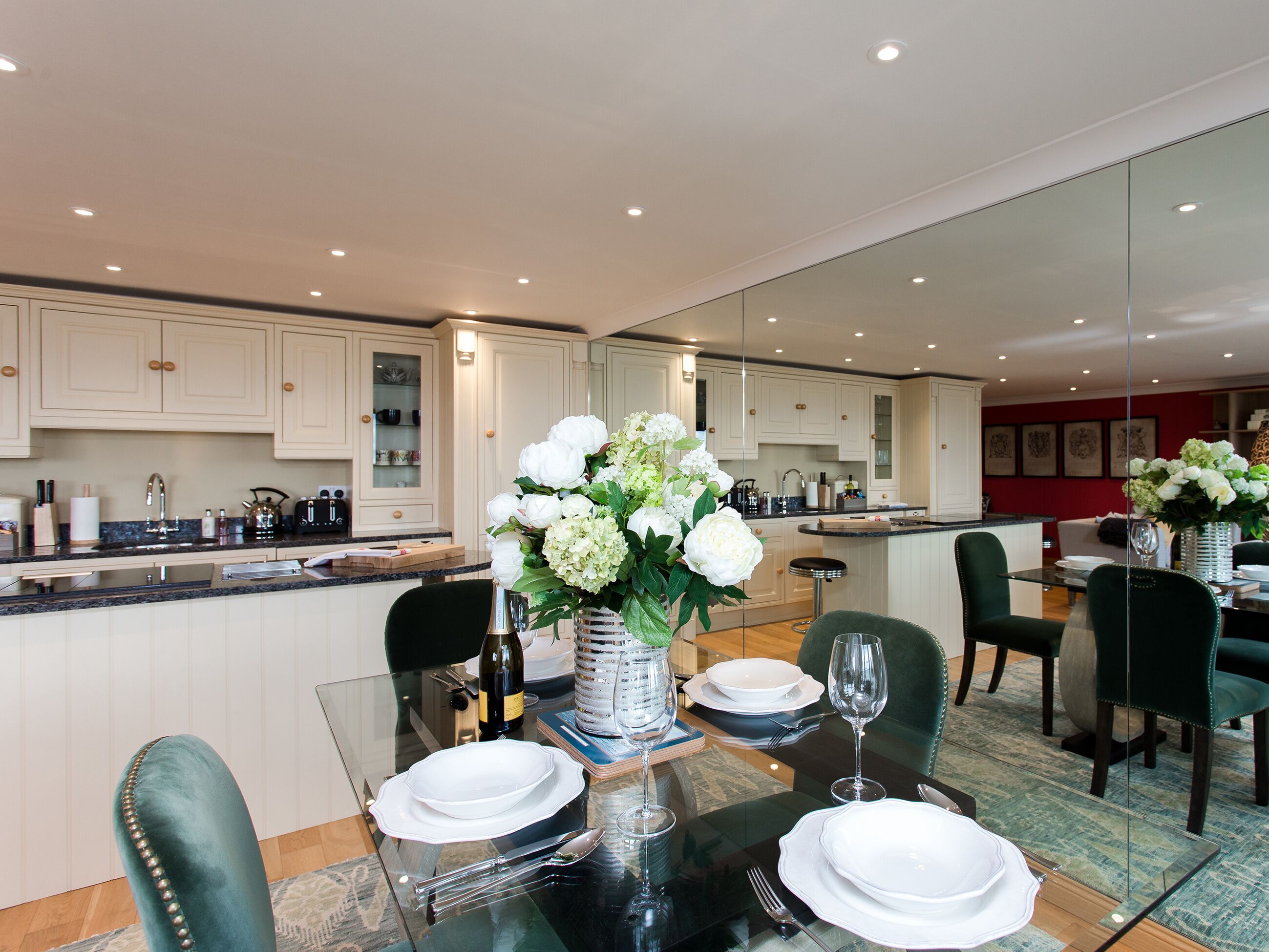 A modern kitchen with white cabinets, a dining area with a glass table, and a large mirror reflecting the space.