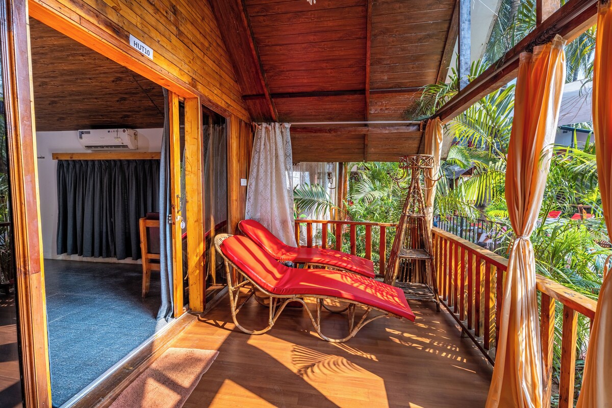 A wooden balcony with two red lounge chairs, a wooden railing, and a view of lush greenery.