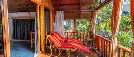 A wooden balcony with two red lounge chairs, a wooden railing, and a view of lush greenery.