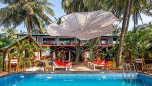 A pool area with red lounge chairs, a large umbrella, and a building with balconies.