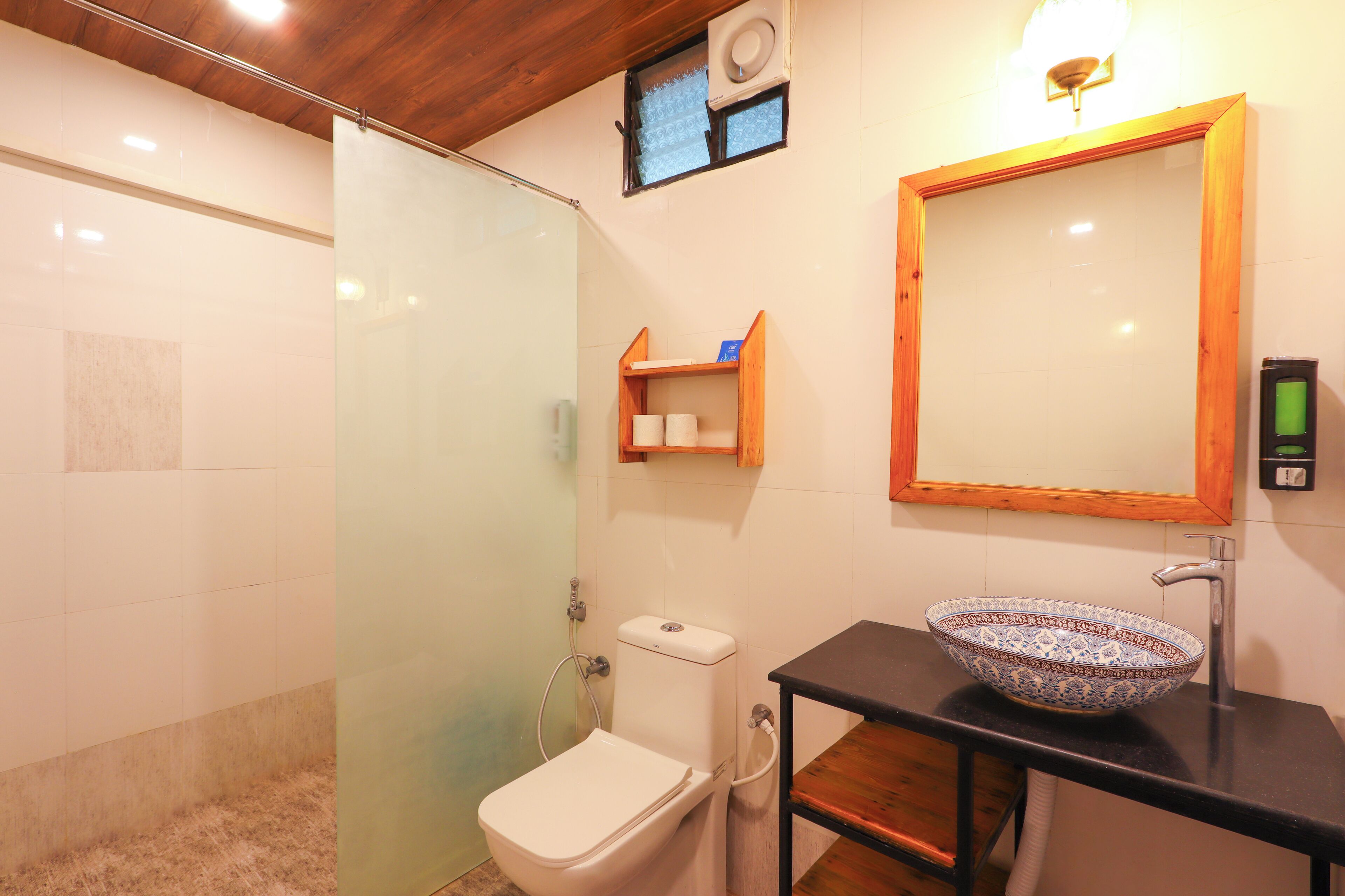 A bathroom with a glass shower enclosure, a toilet, a sink with a decorative bowl, a wooden shelf with toiletries, and a mirror with a wooden frame.