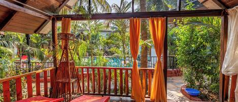 A covered outdoor area with a wooden deck, red lounge chairs, and a view of a pool and palm trees.