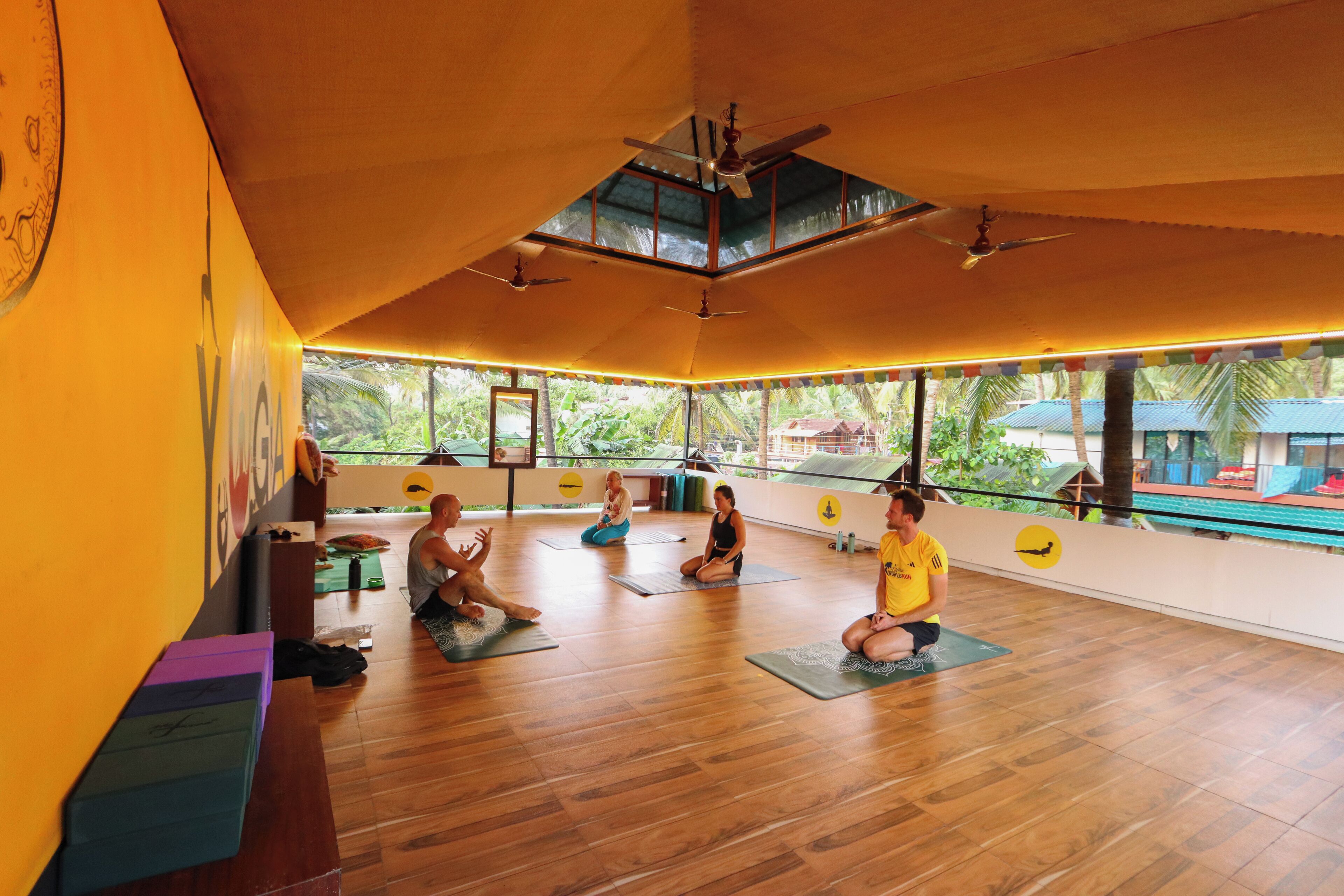 Un studio de yoga doté de planchers en bois, de ventilateurs de plafond et de grandes fenêtres offrant une vue sur des palmiers et une piscine.