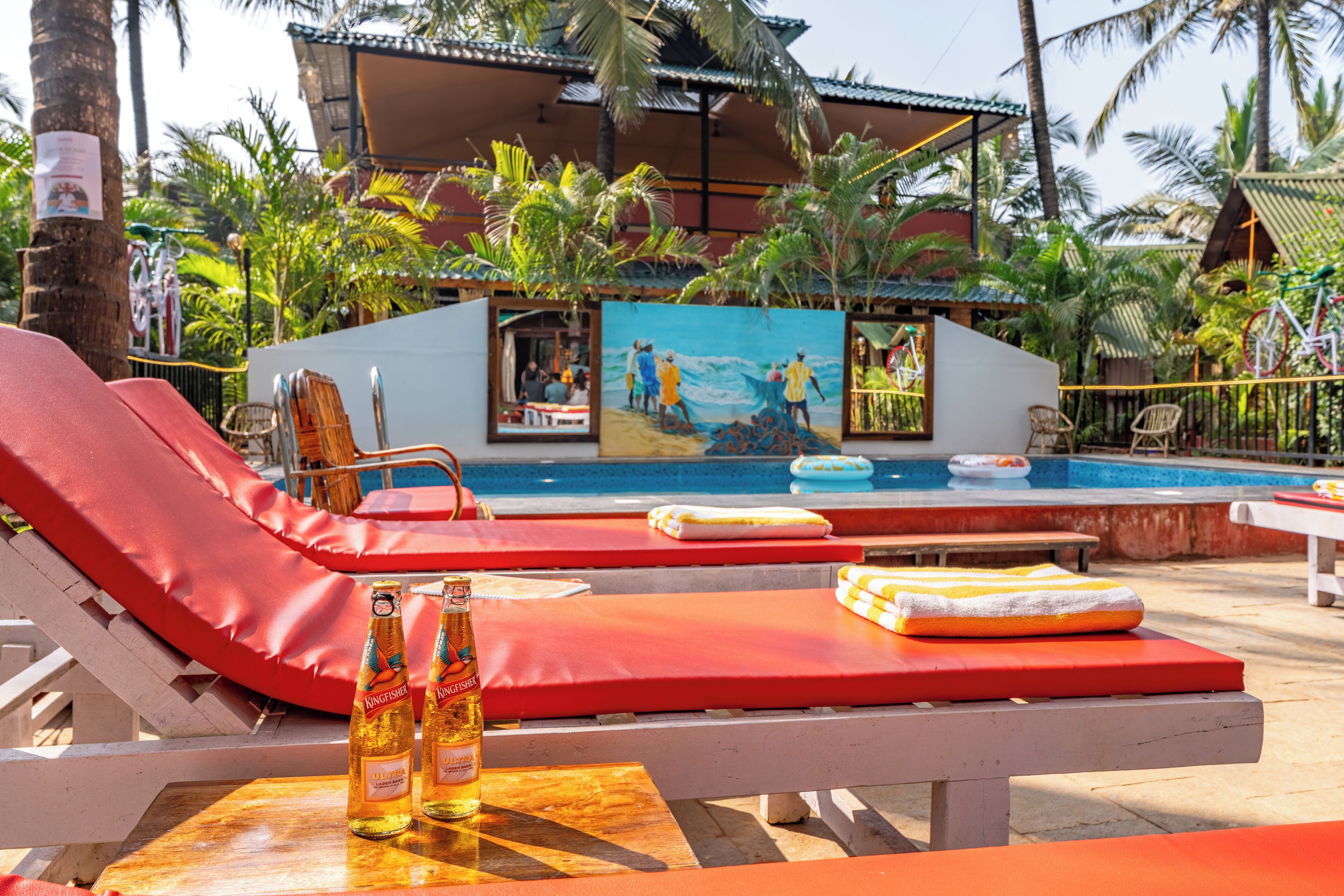 Un espace aménagé au bord de la piscine, avec des chaises longues, une table sur laquelle sont posées des bouteilles, et une fresque représentant des scènes de plage.
