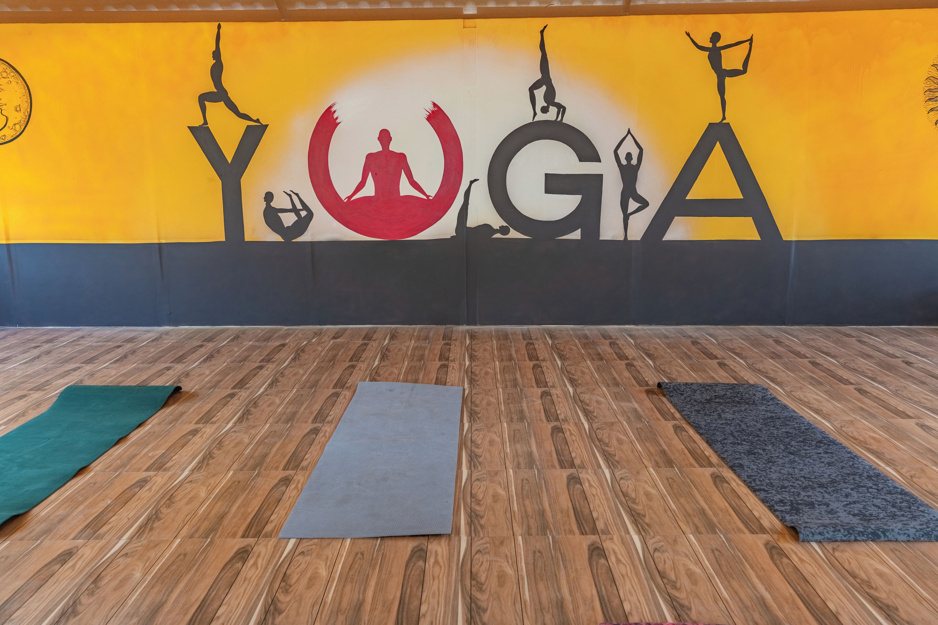 A yoga studio with a wooden floor, a large wall mural with the word "YOGA" and silhouettes of yoga poses, and two yoga mats.