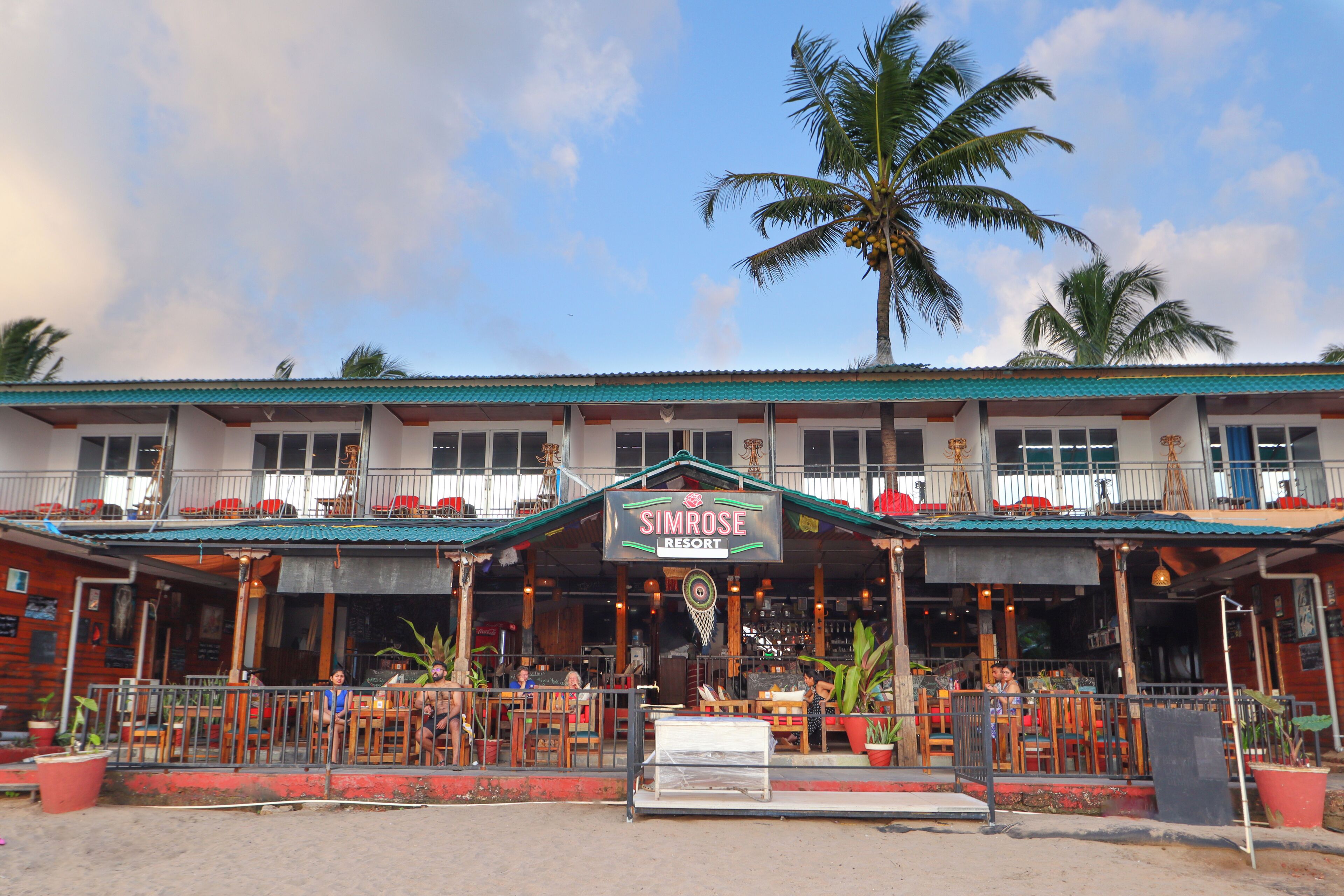 A two-story building with a sign reading "SIMROSE RESORT." There are outdoor seating areas with tables and chairs, and a palm tree is visible in the background.