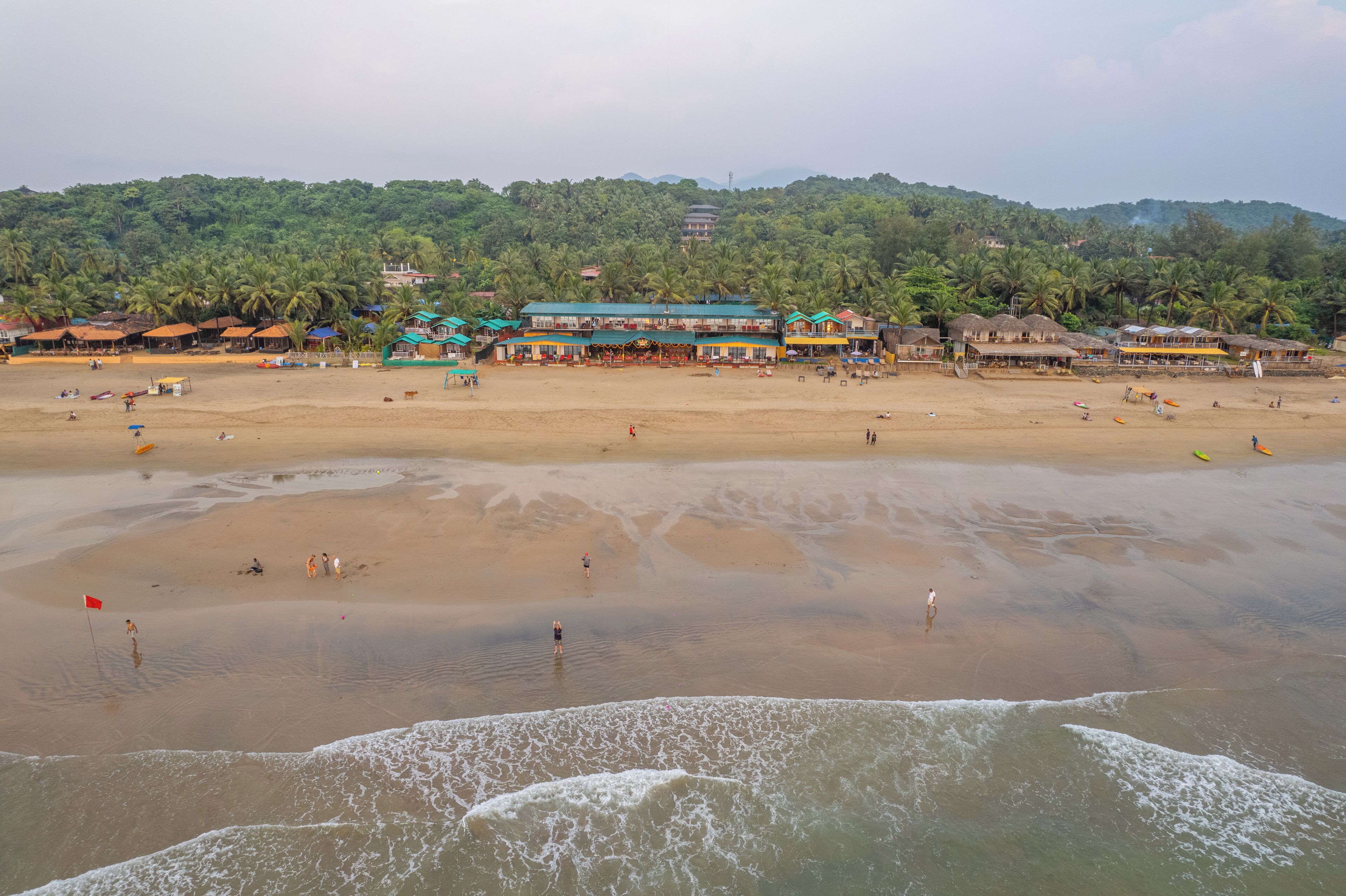 A beach with a row of buildings, some with thatched roofs, and a forested hill in the background.