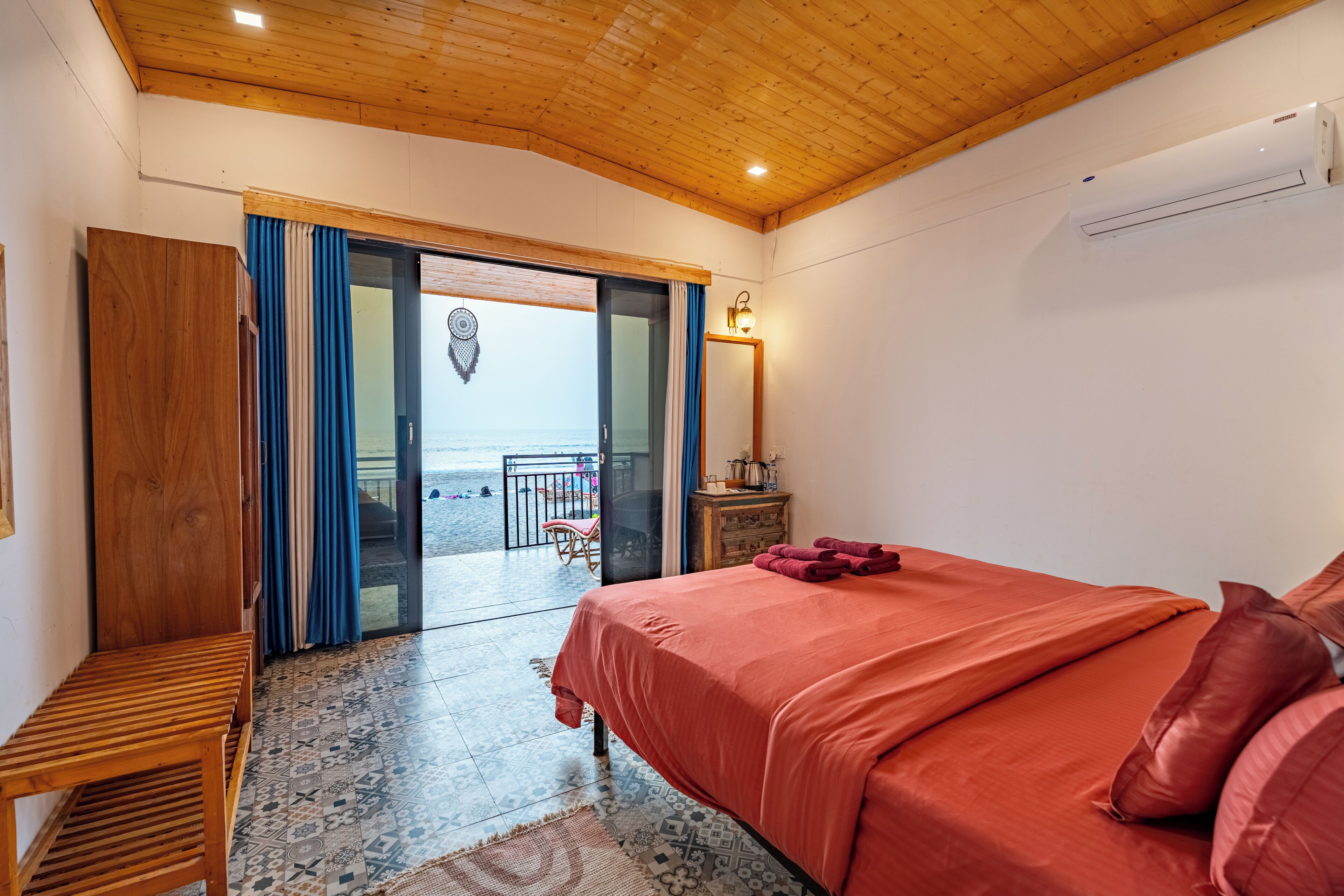 Ein Schlafzimmer mit Holzbalkendecke, einem Bett mit roter Bettwäsche, einer hölzernen Bank und Blick auf den Strand durch eine geöffnete Tür.