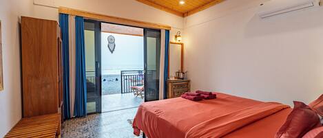 Ein Schlafzimmer mit Holzbalkendecke, einem Bett mit roter Bettwäsche, einer hölzernen Bank und Blick auf den Strand durch eine geöffnete Tür.