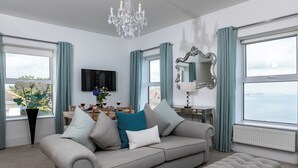 Un salon avec un canapé gris, un pouf assorti, un lustre, une table à manger avec des chaises, un téléviseur et une vue sur la mer grâce à de grandes fenêtres.