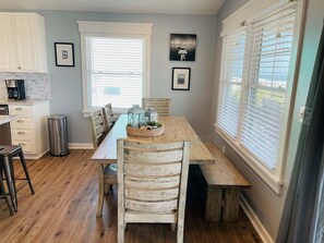 Un coin repas avec une table en bois, des chaises et un banc. Il y a deux fenêtres avec des stores, une poubelle et des tableaux encadrés au mur.