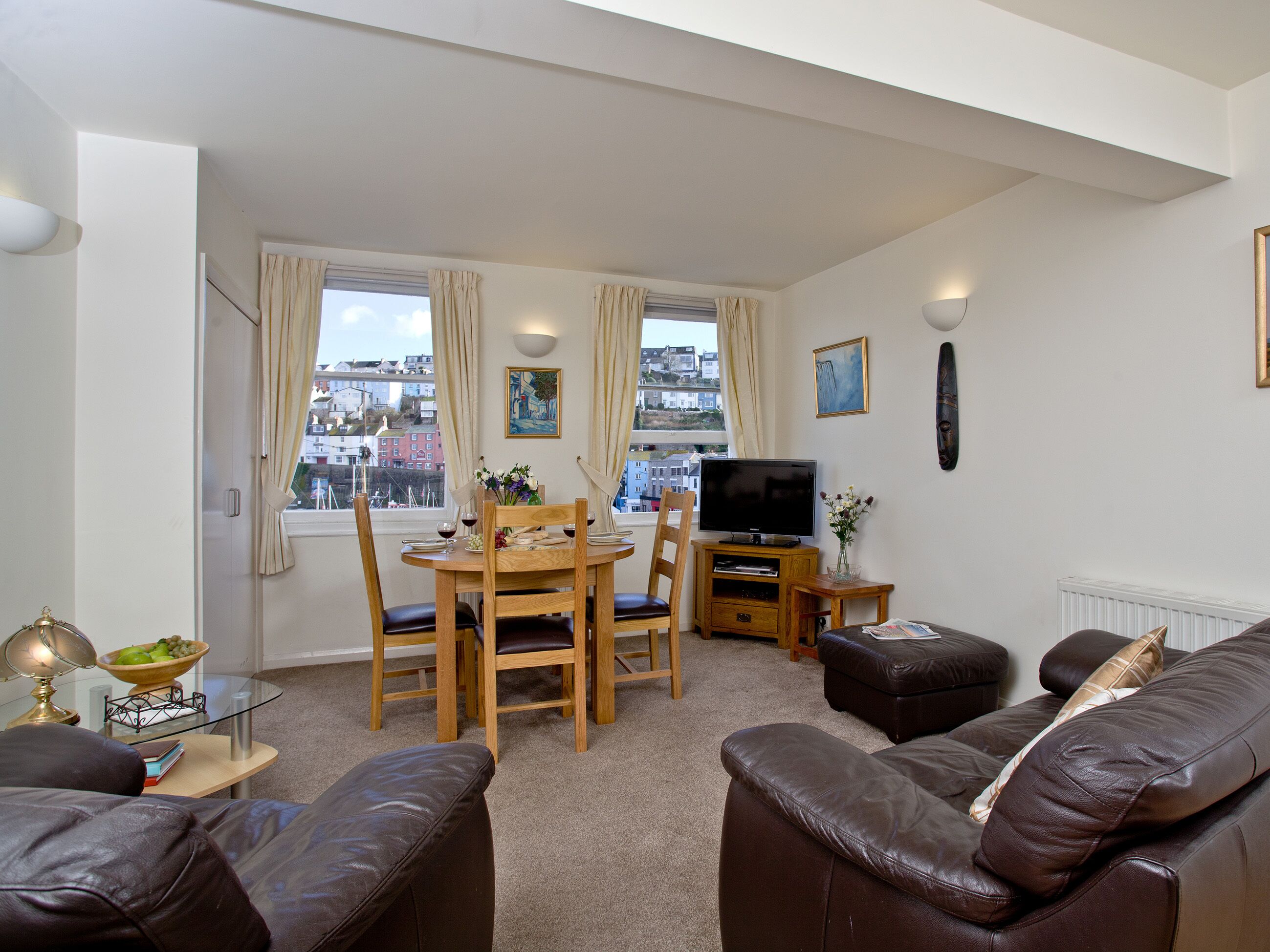 A living room with a wooden dining table, chairs, a television, a sofa, and a view of buildings through the windows.