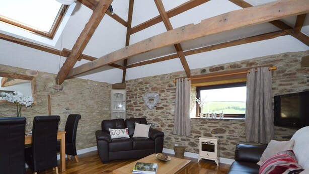A cozy living room with a stone wall, wooden beams, a black leather sofa, a wooden coffee table, and a television.