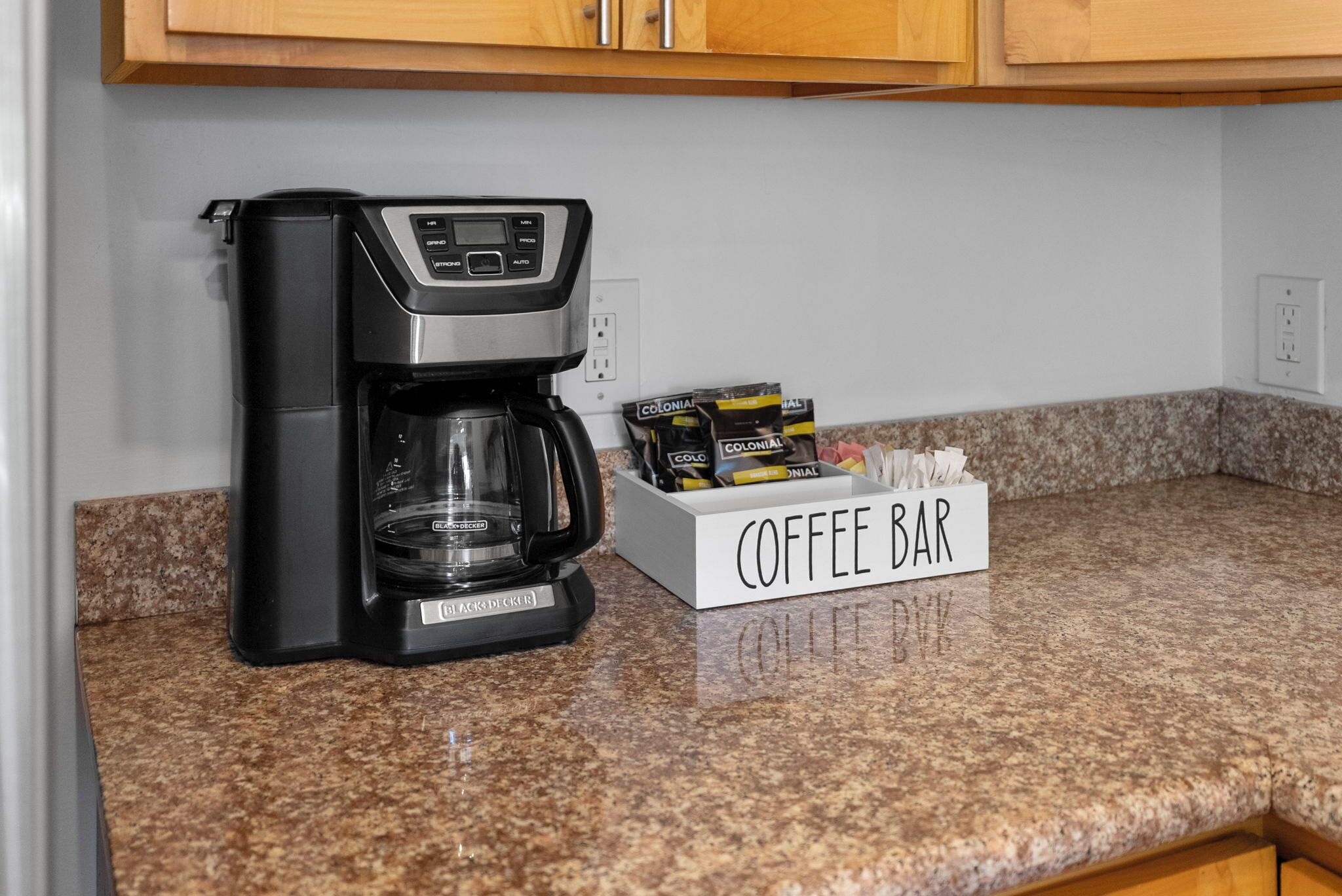 Una cafetera sobre una encimera de granito con un cartel de cafetería y cápsulas de café.
