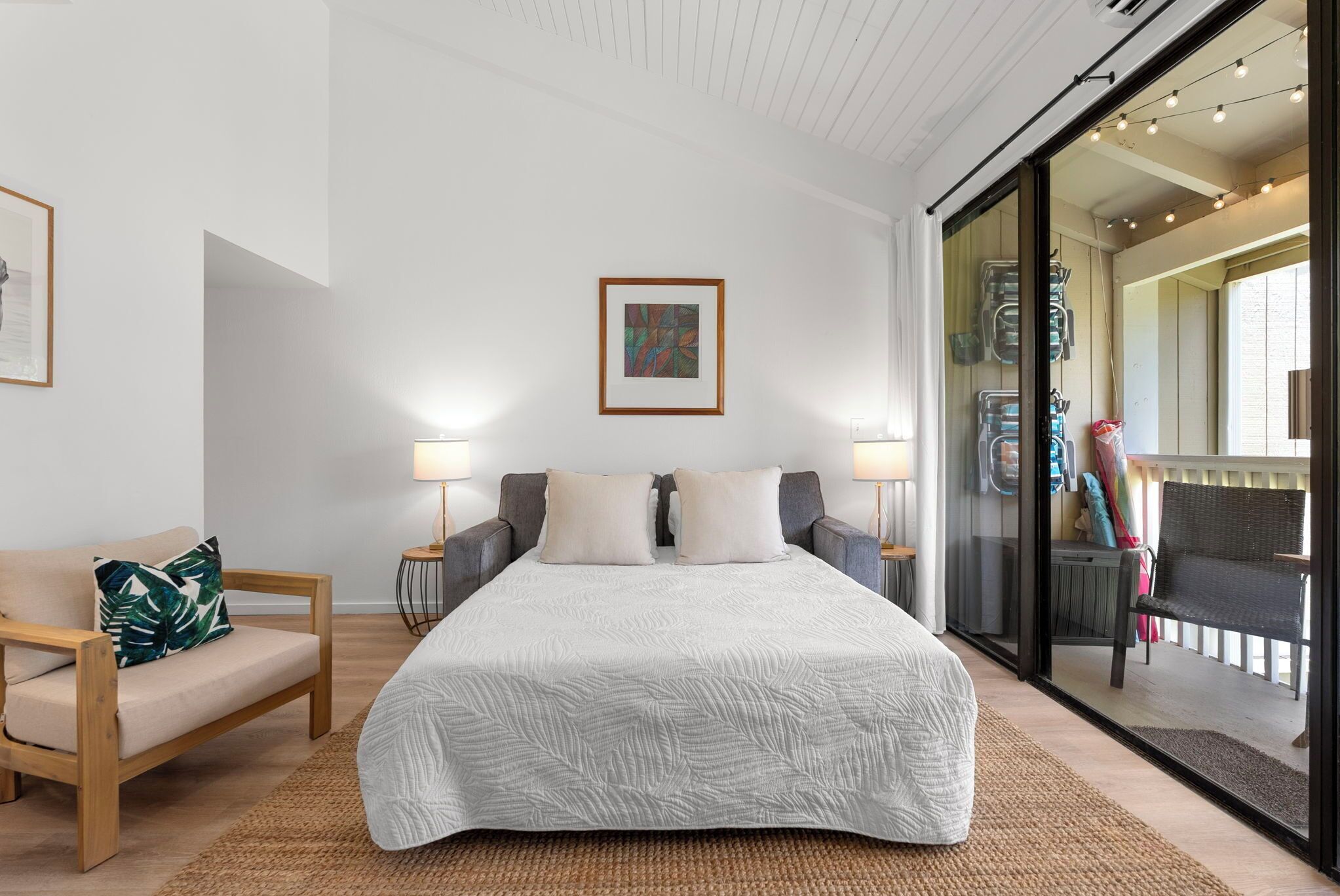 A bedroom with a large bed, two bedside tables with lamps, a wooden chair, and a view through a glass door to an outdoor area.