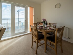 A dining area with a table set for four, a clock on the wall, and a view of the sea through large windows. - 2 bedroom accommodation in Shanklin (Shanklin)